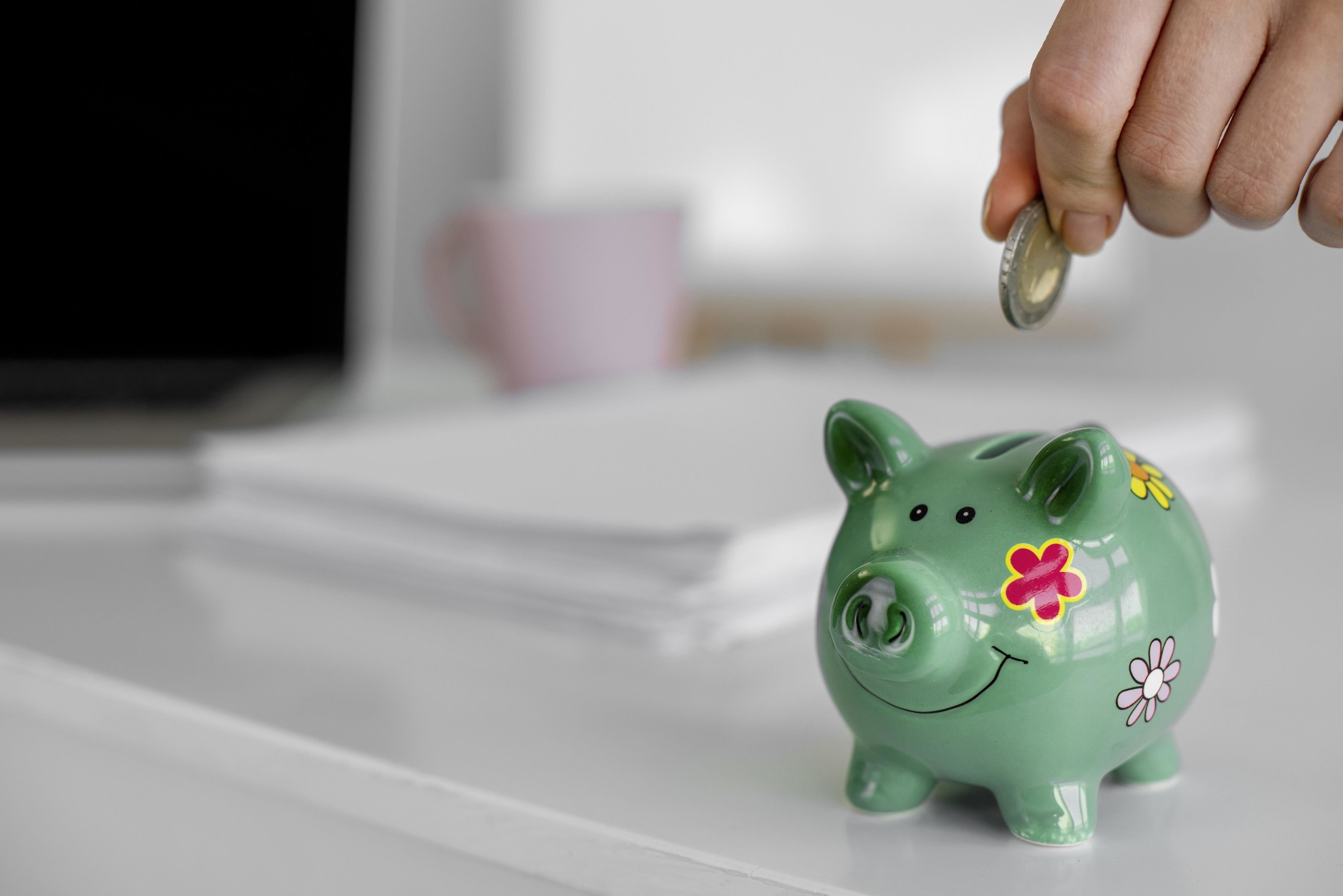 Una hucha en forma de cerdito de color verde. La mano de una persona acerca una moneda para meterla dentro